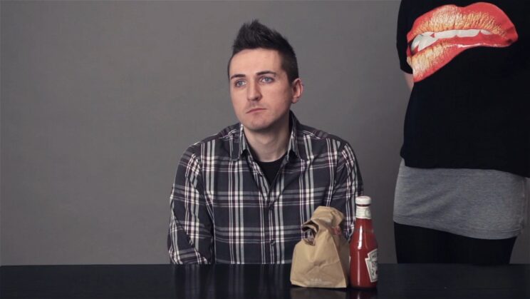 The artist is sitting behind a table with a bag of hamburgers in front of him. A woman is standing next to the table. Her T-shirt has a picture of a woman's mouth on it.